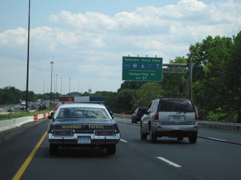 1991 Ford LTD Crown Victoria - black sedan lettered HIGHWAY PATROL drives next to a silver SUV beneath a green sign for the Baltimore Travel Plaza and Canton/Fells Pt exi…