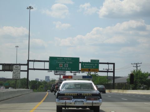 1991 Ford LTD Crown Victoria - black-and-white HIGHWAY PATROL cruiser leads traffic beneath green overhead signs for Inner Harbor, Camden Yards and the O'Donnell St exit.