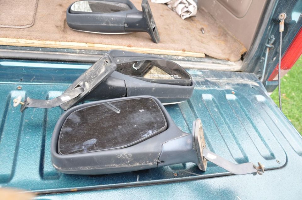 Two black side mirrors, one with a cracked glass, lying in the bed of a teal truck