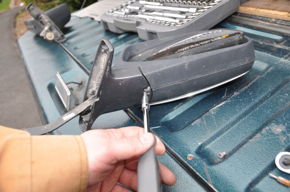 Black car side mirror with chrome trim, being worked on with a screwdriver on a green truck bed