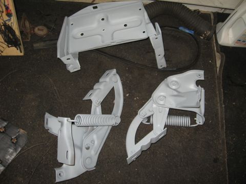 1966 Ford F-100 - Two gray hood hinges with springs and a gray metal bracket are laid out on a dark surface.