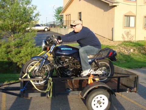 1985 Ford LTD Crown Victoria - A man sits on a Suzuki motorcycle being transported on a trailer.