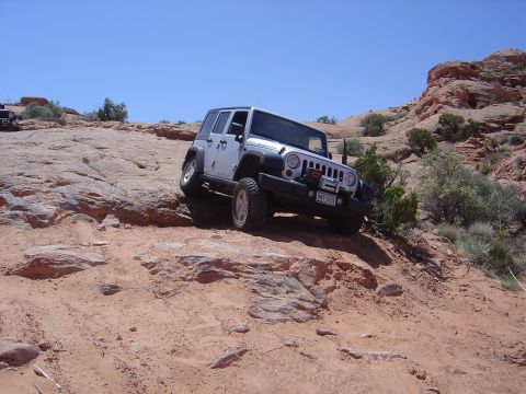 1986 Ford Bronco II - A Jeep navigates a challenging off-road trail with red rock formations and sparse vegetation under a clear blue sky.