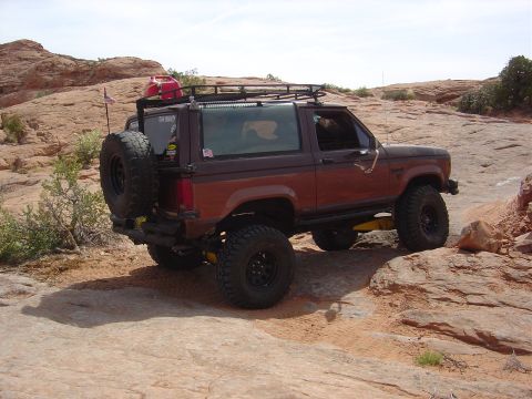 1986 Ford Bronco II - A brown Ford Bronco II navigates a rocky off-road path, equipped with a spare tire and roof rack.