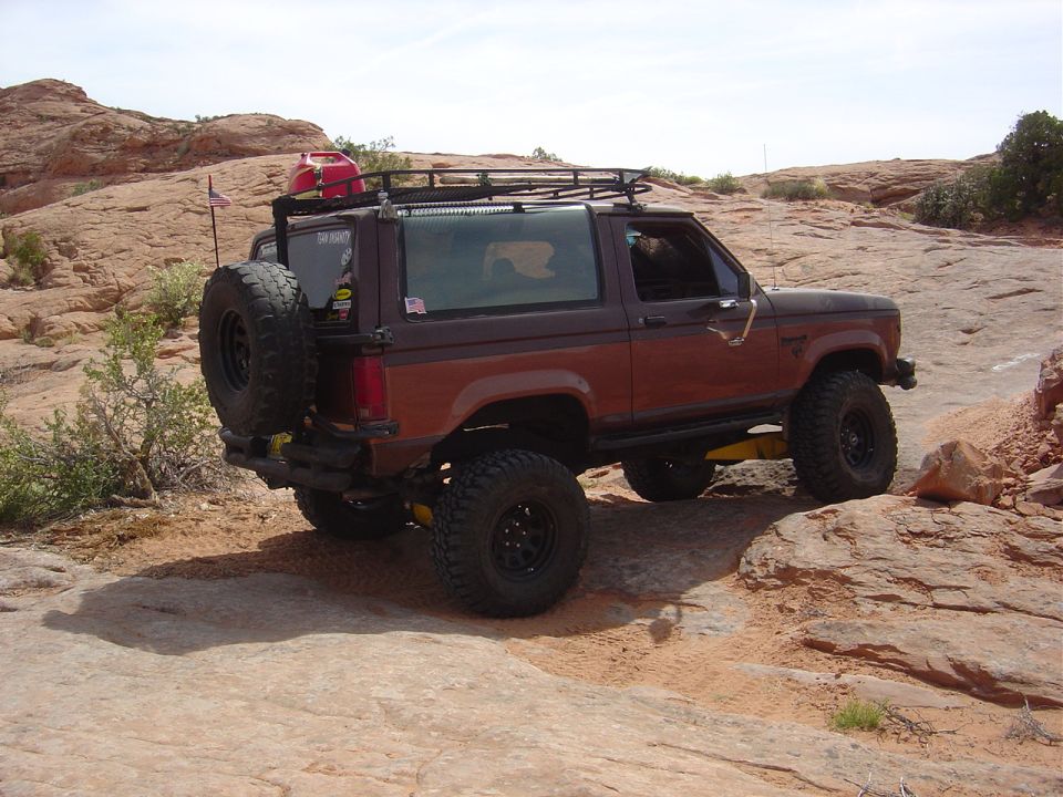 Brown Ford Bronco II with a spare tire on the back, roof rack, and red gas can, driving on a rocky trail