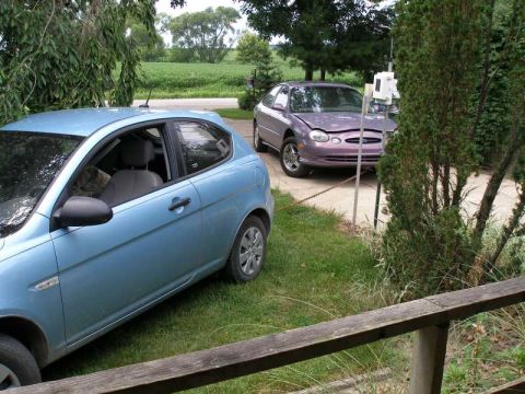 1997 Ford Taurus - light blue Hyundai Accent hatchback parked beside a purple Ford Taurus sedan in a rural driveway by farm fields.