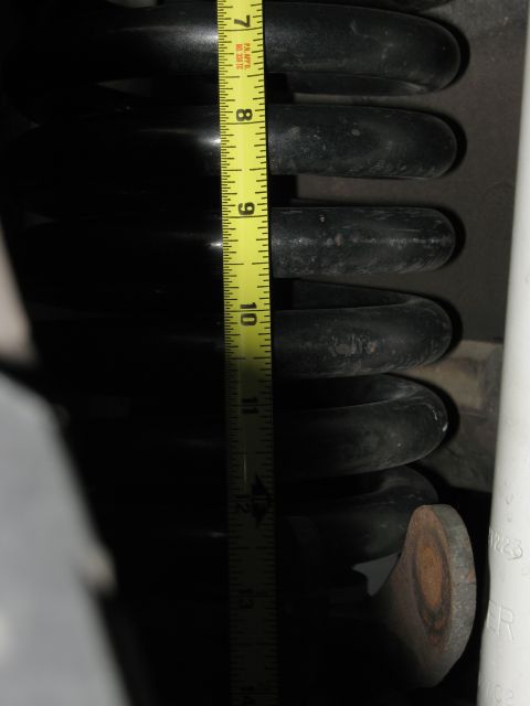 1986 Ford Bronco - A close-up view of a black coil spring being measured with a yellow tape measure, indicating its size.