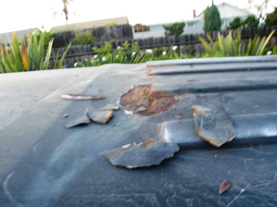 Close-up of a dark-colored vehicle roof with significant rust damage and peeling paint. Background shows greenery and a fence