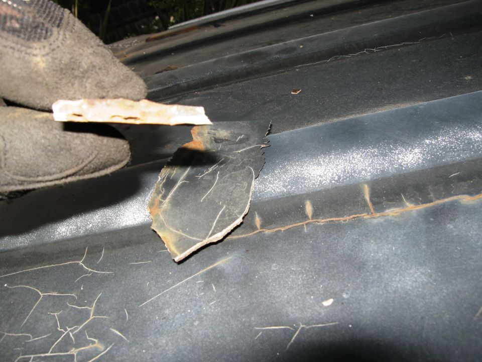 Gloved hand peeling paint from dark vehicle roof showing rust and cracking