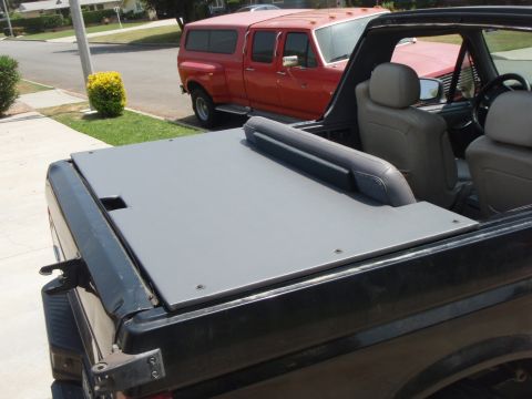 1995 Ford Bronco - Rear view looking forward across the back of a dark-colored convertible/soft-top vehicle with the top down, showing a gray padded tonneau o…