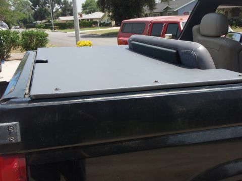 1995 Ford Bronco - Close-up of a black flat tonneau-style cover with visible bolt fasteners over the bed of a dark-colored truck, with a black seat/headrest v…