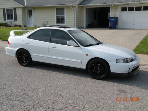 1994 Acura Integra - A white 1994 Acura Integra sedan is parked in front of a suburban home, aftermarket black wheels and a rear spoiler.