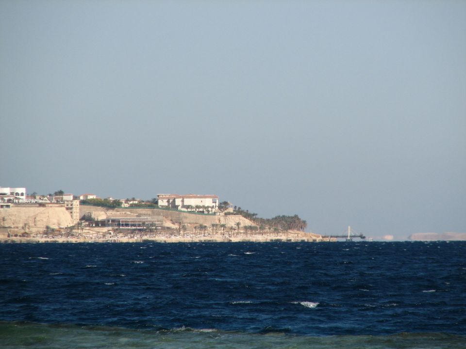 Choppy blue sea with a distant rocky coastline, resort buildings, palm trees, and a pier under hazy sky