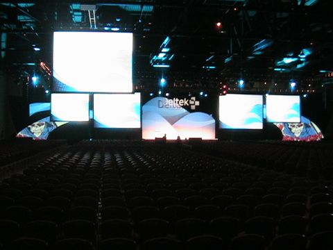 1966 Ford Bronco - A large conference hall stage features multiple screens displaying graphics and the Deltek logo, with rows of empty seats visible.