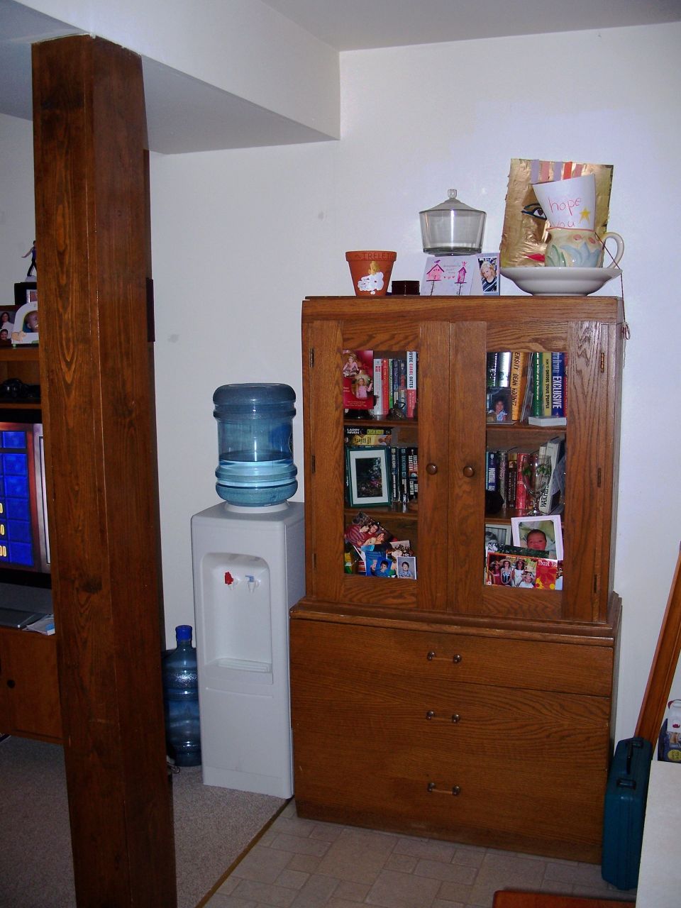 Room interior showing a wooden cabinet filled with books and photos, a white water cooler with a blue water jug, and a large wooden pillar