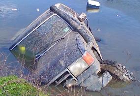 1990 Ford Bronco - A Ford Bronco is seen partially submerged in water, tilted on its side near a grassy bank.