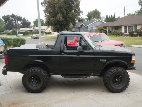 1995 Ford Bronco - A black Ford Bronco with the top removed, shown in side profile with a lift kit and off-road tires on black steel wheels, parked on a drive…