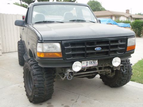 1995 Ford Bronco - Front three-quarter view of a lifted black OBS Ford Bronco with the top removed, sitting on aggressive mud-terrain tires in a residential d…