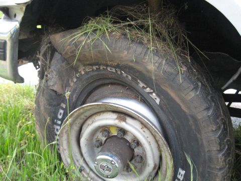 1995 Ford C-350 (Centurion Conversions) - A close-up view of a damaged front wheel and tire, with grass growing around it, suggesting the vehicle is not in use.