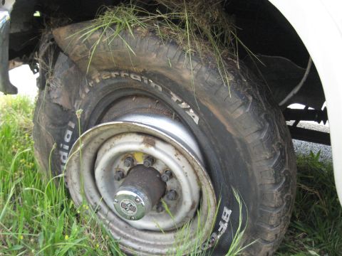 1995 Ford C-350 (Centurion Conversions) - Worn tire and rim detail, likely from an older truck or SUV, overgrown with grass.
