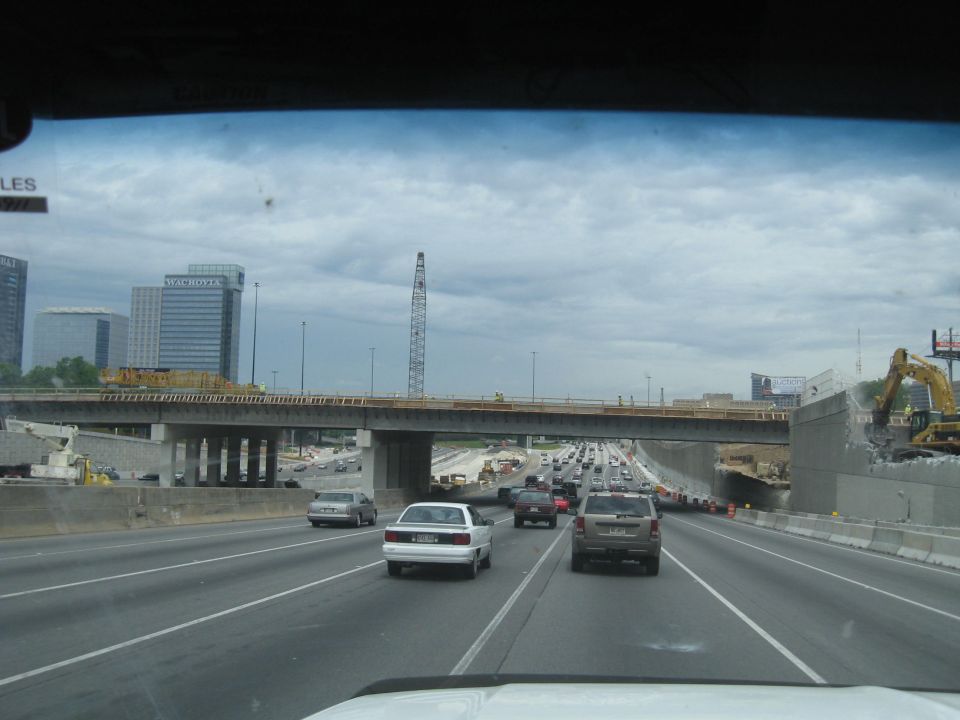 Highway construction with an excavator demolishing a wall, multiple cars on the road, and buildings in the background