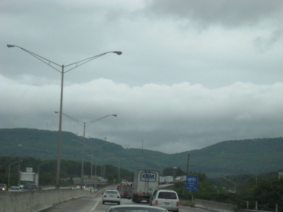 Highway traffic jam under cloudy sky with mountains in background. Streetlights line the road