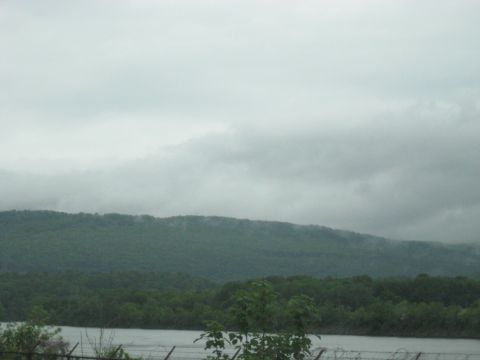 1995 Ford C-350 (Centurion Conversions) - A hazy, overcast sky looms above dense green trees and a calm body of water, with a barbed wire fence visible.