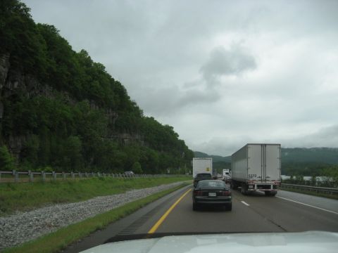 1995 Ford C-350 (Centurion Conversions) - Vehicles travel on a highway under a cloudy sky, with a steep, tree-covered cliff face to the left.