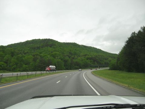 1995 Ford C-350 (Centurion Conversions) - A tanker truck travels down a highway past a wooded hillside on an overcast day.