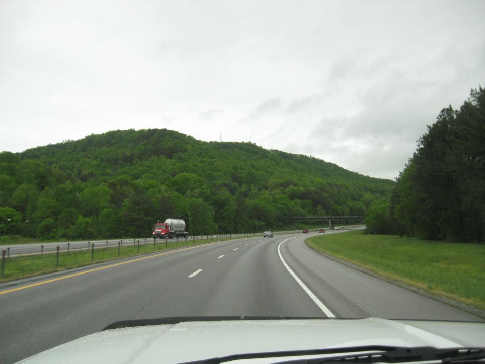 View from a car on a highway, passing a tanker truck. Green, tree-covered hills in the background under a cloudy sky