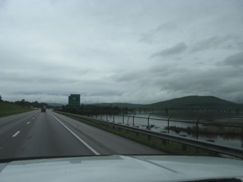 1995 Ford C-350 (Centurion Conversions) - A highway scene unfolds under a cloudy sky, with a green directional sign visible in the distance and a body of water bordering the road.