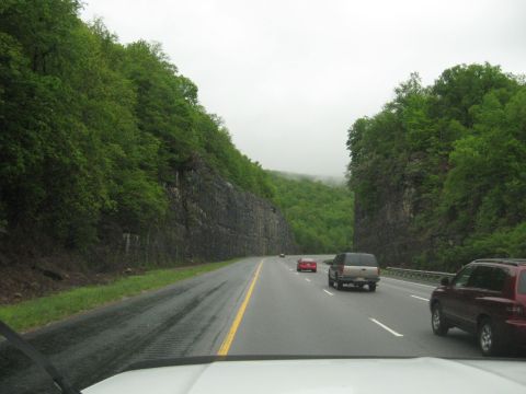1995 Ford C-350 (Centurion Conversions) - Vehicles travel on a wet highway surrounded by lush green trees and rocky cliffs.
