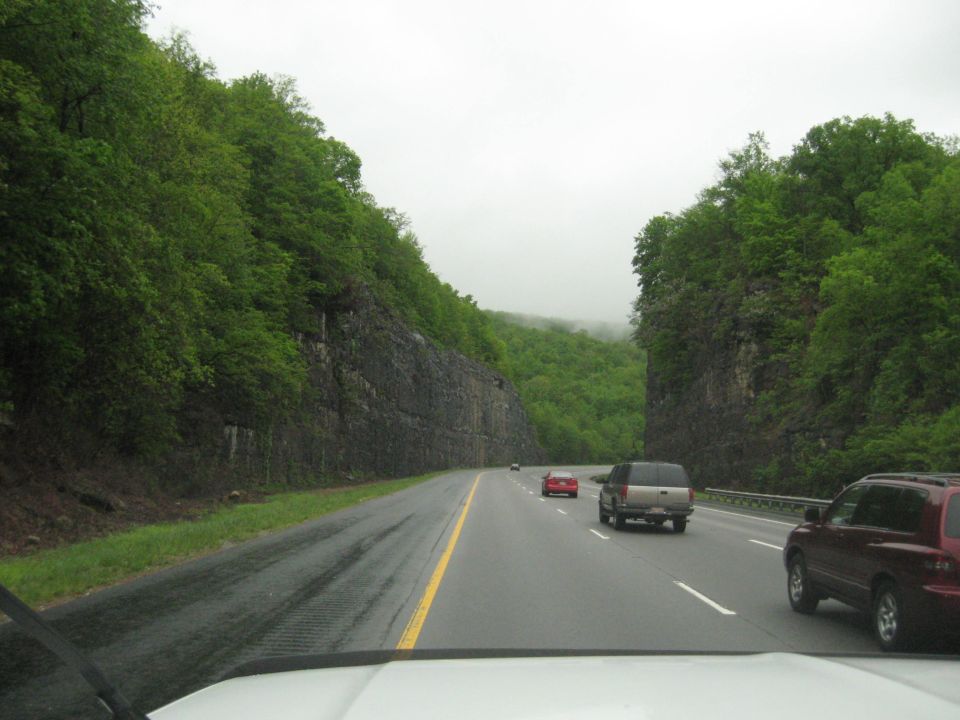 Driving on a wet highway through a forested mountain pass with several vehicles ahead