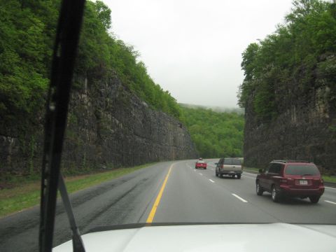 1995 Ford C-350 (Centurion Conversions) - Vehicles travel on a wet highway bordered by steep, tree-covered cliffs under an overcast sky.