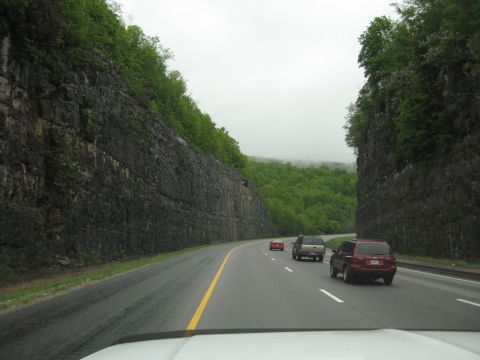 1995 Ford C-350 (Centurion Conversions) - Vehicles travel on a highway flanked by sheer rock cliffs and lush green trees under an overcast sky.