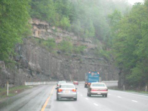 1995 Ford C-350 (Centurion Conversions) - Multiple vehicles travel on a wet highway beside a wooded cliff face.