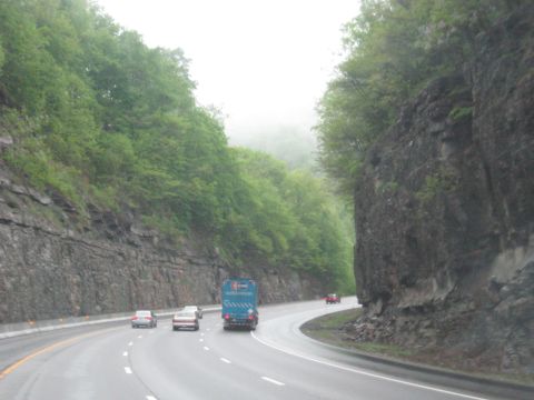 1995 Ford C-350 (Centurion Conversions) - Vehicles travel on a wet highway bordered by steep, tree-covered cliffs.