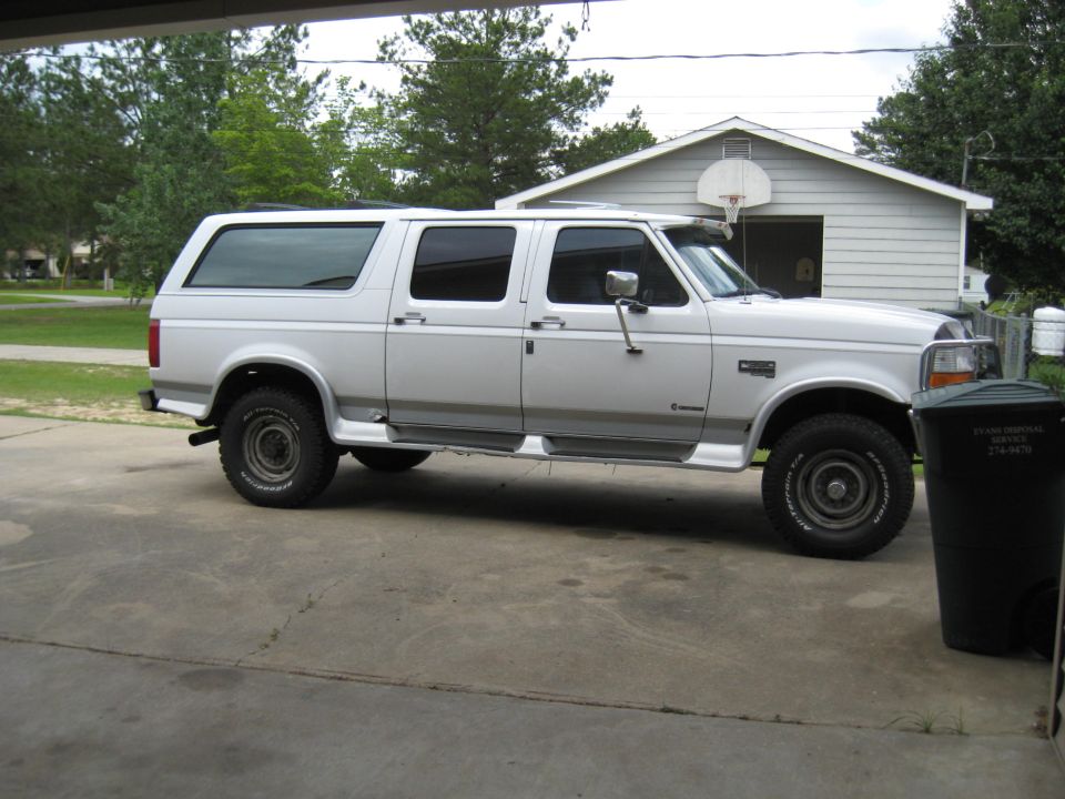 White extended cab pickup truck with a camper shell, side view, parked on concrete. Visible text on trash can