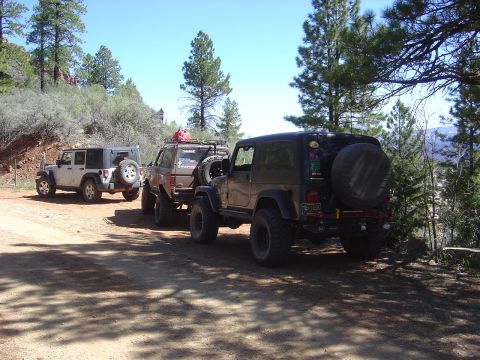 1986 Ford Bronco II - Several off-road vehicles are parked on a dusty trail surrounded by trees and hills under a clear blue sky.