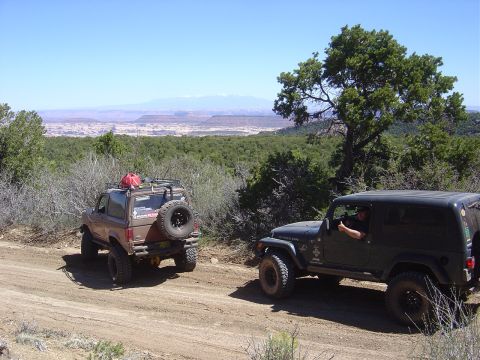 1986 Ford Bronco II - Two off-road vehicles navigate a dusty trail with a vast landscape in the background.