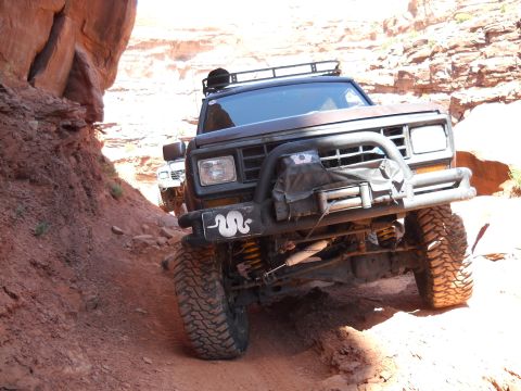1986 Ford Bronco II - Two off-road vehicles tackle a narrow, rocky path in a desert canyon.