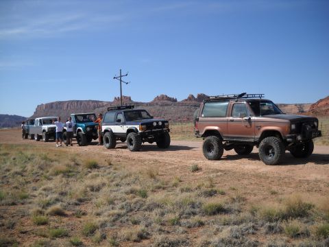 1986 Ford Bronco II - A group of off-road vehicles are lined up on a dirt road with mesas in the background under a clear blue sky.
