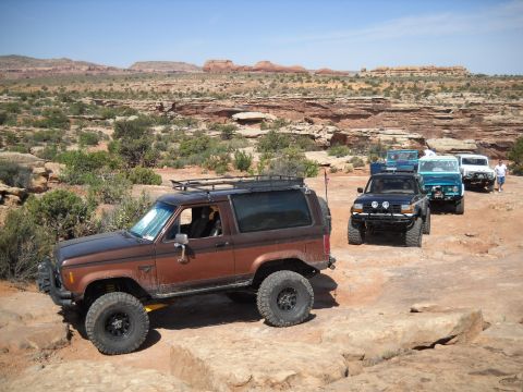 1986 Ford Bronco II - A convoy of off-road vehicles navigates a desert landscape under a clear blue sky.