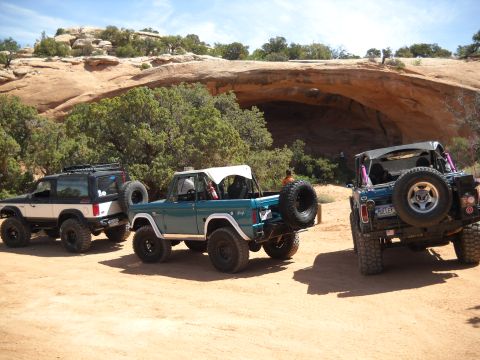 1986 Ford Bronco II - Three modified Broncos are parked in a desert landscape with a large rock overhang.