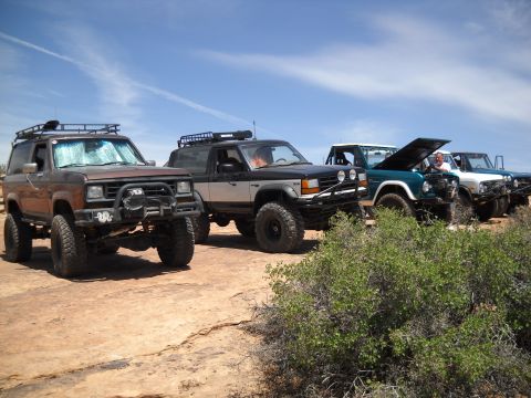 1986 Ford Bronco II - A line of off-road vehicles are parked on a dusty trail with sparse vegetation and a bright blue sky overhead.