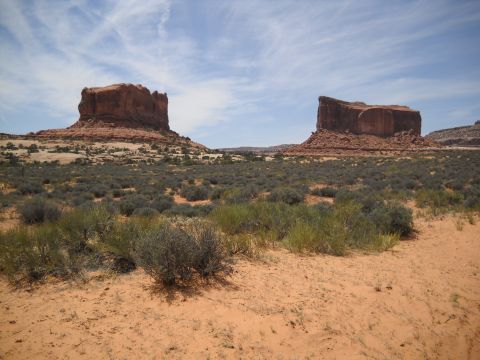 1986 Ford Bronco II - A wide desert scene features towering red rock formations under a partly cloudy sky, with scrub brush and sand in the foreground.