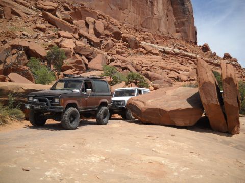 1986 Ford Bronco II - Two off-road vehicles are parked on a dusty trail surrounded by massive red rock formations.