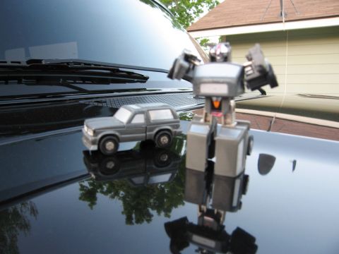 1986 Ford Bronco - A toy robot stands beside a toy SUV, both reflecting on the glossy surface of a vehicle.