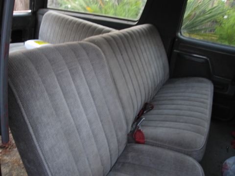 1986 Ford Bronco - The interior of a vehicle shows two rows of gray fabric seats with a red seatbelt buckle.