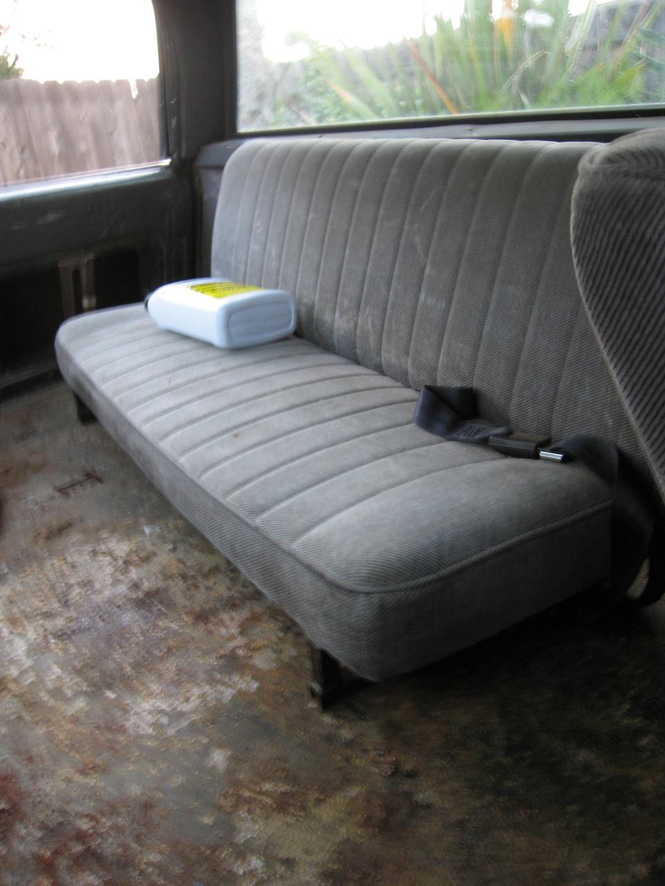Gray fabric bench seat with seatbelt and a yellow-labeled bottle on it. Interior of a vehicle